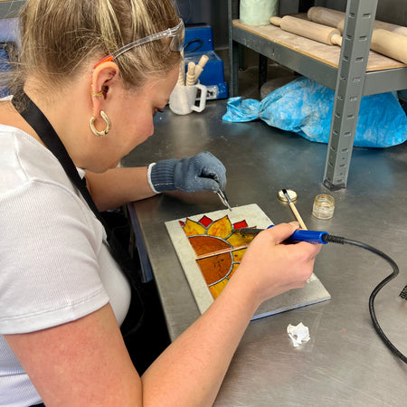 A woman wearing safety glasses and a black apron works on a stained glass piece on a stainless steel table in a workshop setting.