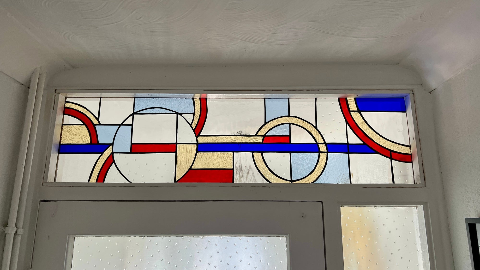 A decorative stained glass window panel with geometric shapes in blue, red, cream, and light blue, installed above a white door.