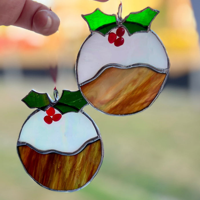 Two stained glass pudding ornaments hanging in sunlight, with a hand holding one of them. The ornaments feature white frosting, brown pastry, green holly leaves, and red berries.