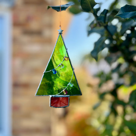 A triangular stained glass Christmas tree ornament hanging outdoors among green leaves, with a blurred brick wall and sky in the background.
