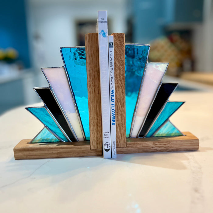 A modern geometric bookend made of light oak wood and translucent blue, white, and black glass triangles, holding two books on a white marble countertop with a blurred kitchen background.