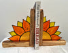 A pair of rustic wooden bookends decorated with hand-painted sunflowers, holding up two books on a white fabric surface against a plain light gray background.