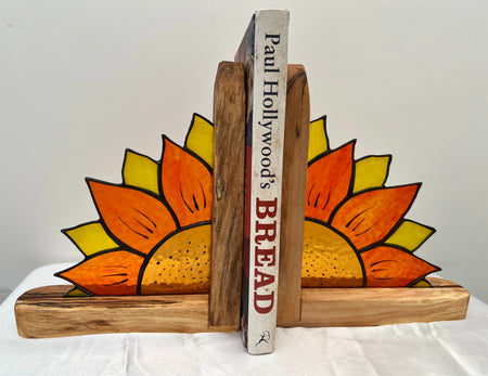 A pair of rustic wooden bookends decorated with hand-painted sunflowers, holding up two books on a white fabric surface against a plain light gray background.