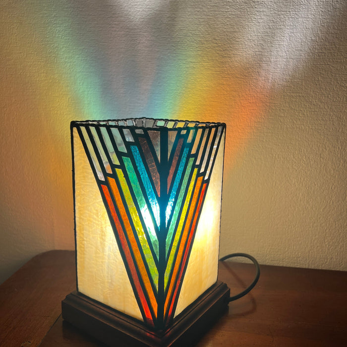 A decorative table lamp with a geometric stained-glass style shade in orange, yellow, green, blue, and red, set on a wooden table against a textured wall. The lamp is turned on, casting a rainbow-colored light pattern onto the wall behind it.