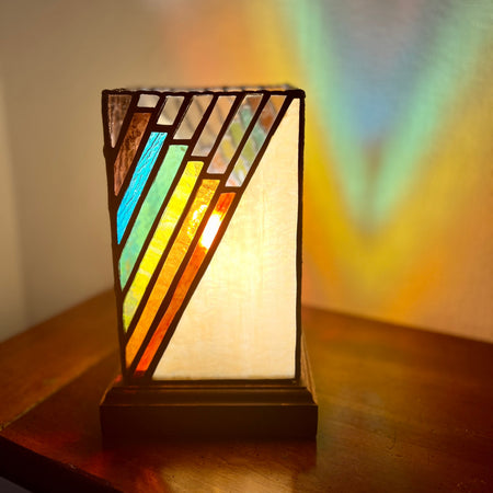 A stained glass table lamp with geometric triangular panels in blue, green, yellow, orange, and white, set on a dark wooden table with warm light projected onto the wall behind it.