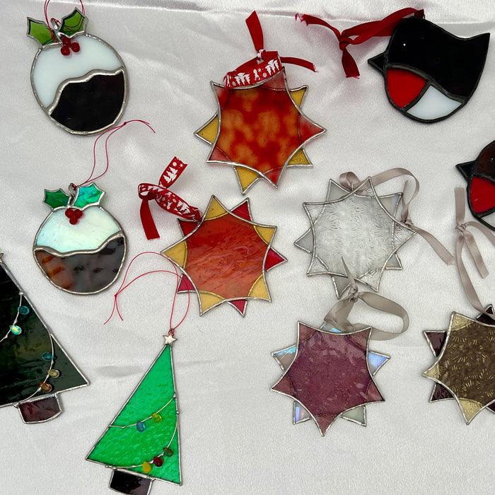 A collection of stained glass Christmas ornaments laid out on a white fabric background, including pudding, penguin, Christmas tree, and star shapes in various colors and textures.