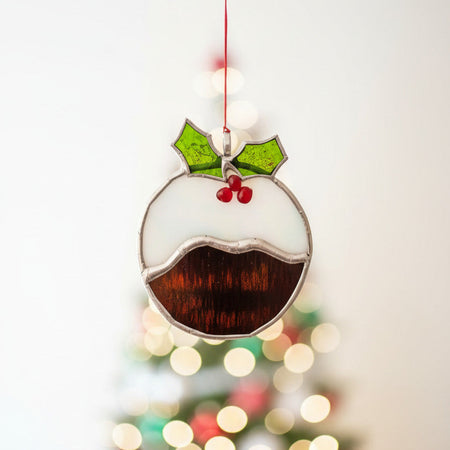 A stained glass Christmas pudding ornament hanging from a thin red string against a plain white wall. The ornament features a black body representing pudding, a white upper section, green holly leaves, and red berries outlined in silver-colored metal.