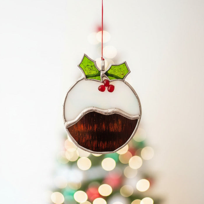 A stained glass Christmas pudding ornament hanging from a thin red string against a plain white wall. The ornament features a black body representing pudding, a white upper section, green holly leaves, and red berries outlined in silver-colored metal.
