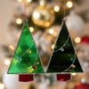 Two stained glass Christmas tree ornaments hanging from a hand against a plain white wall. One ornament is bright green with a silver star top and multicolored glass bead garland, and the other is dark green with a similar garland and a red base piece.