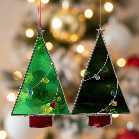 Two stained glass Christmas tree ornaments hanging from a hand against a plain white wall. One ornament is bright green with a silver star top and multicolored glass bead garland, and the other is dark green with a similar garland and a red base piece.