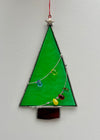 A stained glass Christmas tree ornament with a bright green body, silver metal frame, a silver star topper, and small multicolored glass beads on a wire, hanging against a white wall.
