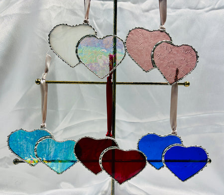 A set of heart-shaped stained glass ornaments in multiple colors, including white, pink, blue, and red, hanging from a metal stand against a white fabric backdrop.