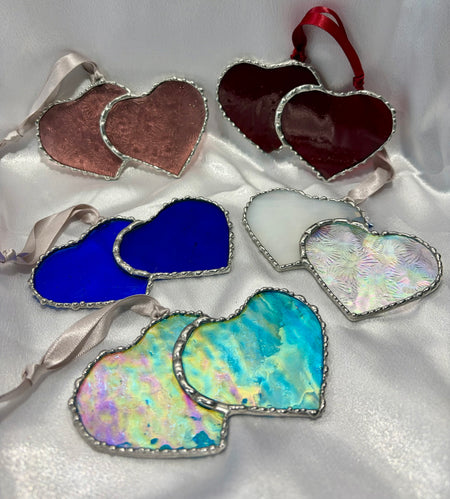 Six heart-shaped stained glass ornaments with silver borders and hanging ribbons, displayed on a white satin background. The ornaments feature various colors and iridescent patterns.
