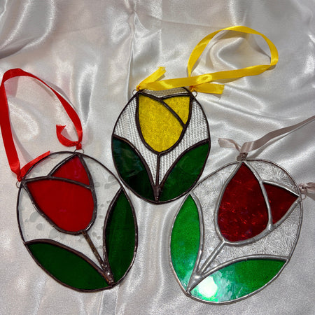 Three tulip-shaped stained glass ornaments with hanging ribbons, displayed on a white satin fabric background. One ornament has a red tulip with green leaves and a red ribbon, while the other two feature yellow and red tulip designs with green leaves and silver-toned metal frames.