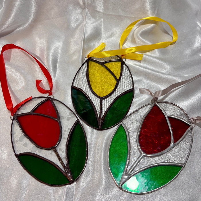 Three tulip-shaped stained glass ornaments with hanging ribbons, displayed on a white satin fabric background. One ornament has a red tulip with green leaves and a red ribbon, while the other two feature yellow and red tulip designs with green leaves and silver-toned metal frames.