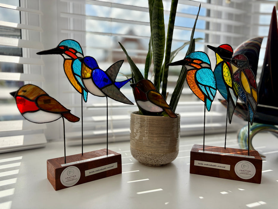 Three stained glass bird sculptures on wooden bases displayed on a windowsill with a potted snake plant in the center. The birds are made of colorful glass panels with black metal wire stands and rectangular wood bases with round white labels.