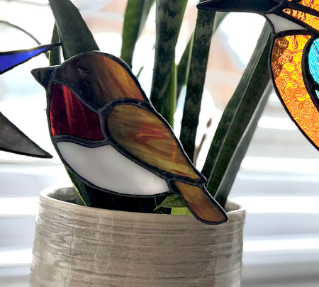 A stained glass robin bird ornament with brown, white, and red sections, displayed in a textured beige pot among green plant leaves near a window.
