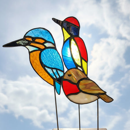 three stained glass bird sculptures on metal stands, featuring colorful bird designs with blue, red, yellow, orange, white, and brown panels separated by dark lead lines, displayed indoors
