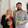 A man and a woman standing indoors, each holding a stained glass heart-shaped ornament with blue, yellow, green, and orange sections. The woman wears a black cardigan over a red floral dress, and the man wears a light patterned button-up shirt.