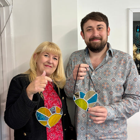 A man and a woman standing indoors, each holding a stained glass heart-shaped ornament with blue, yellow, green, and orange sections. The woman wears a black cardigan over a red floral dress, and the man wears a light patterned button-up shirt.