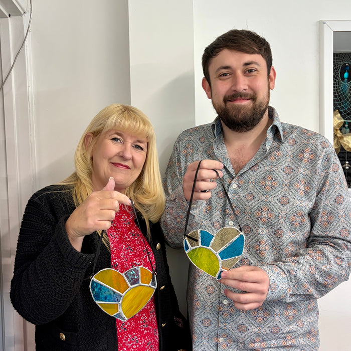 A man and a woman standing indoors, each holding a stained glass heart-shaped ornament with blue, yellow, green, and orange sections. The woman wears a black cardigan over a red floral dress, and the man wears a light patterned button-up shirt.