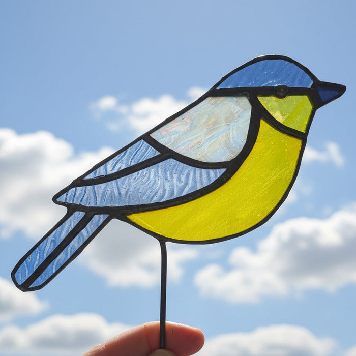 A stained blue tit ornament with a blue and yellow design, held by a hand against a blue sky with white clouds.