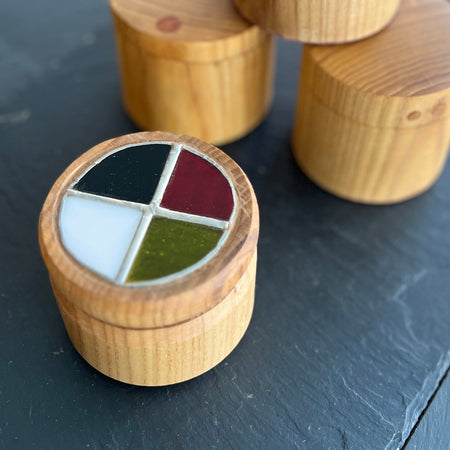 Four small round wooden boxes with lids, arranged on a dark slate surface. One box in the foreground is open and shows a four-quadrant painted lid with black, white, red, and green sections.
