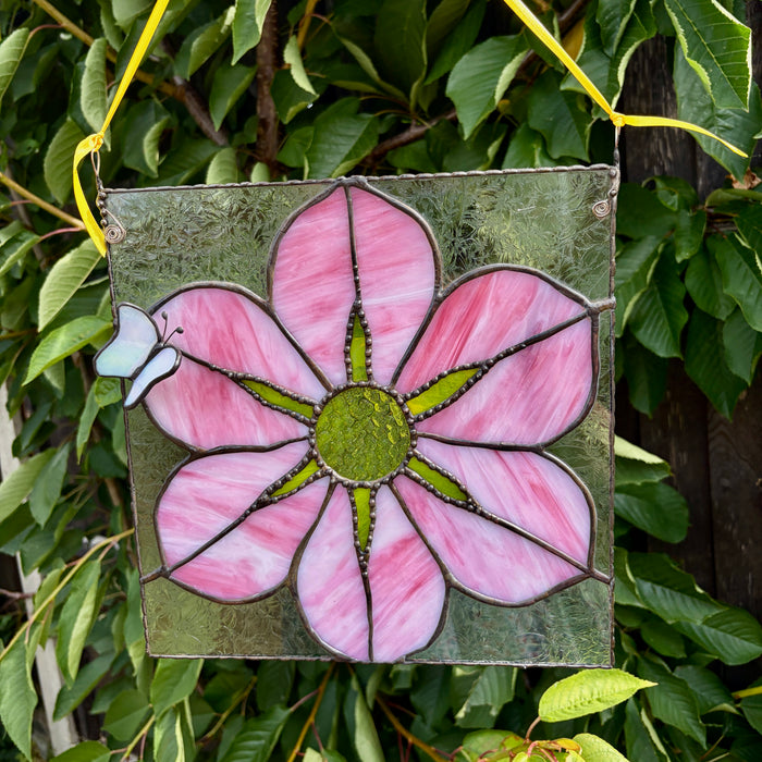 A square stained glass hanging panel featuring a large pink flower with a green center, suspended by a yellow ribbon against a background of green leaves.