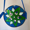 A circular stained glass ornament featuring a green Celtic knot design over blue and white glass panels, suspended by a blue ribbon against a plain light background.