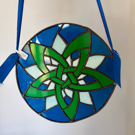 A circular stained glass ornament featuring a green Celtic knot design over blue and white glass panels, suspended by a blue ribbon against a plain light background.