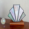 A stained glass art deco lamp shade with iridescent mother-of-pearl panels, mounted on a wooden base, displayed on a wooden table next to a small clear crystal sphere against a textured off-white wall.