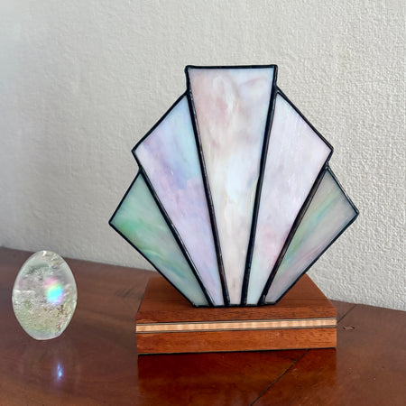 A stained glass art deco lamp shade with iridescent mother-of-pearl panels, mounted on a wooden base, displayed on a wooden table next to a small clear crystal sphere against a textured off-white wall.