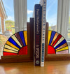 Two wooden bookends with stained-glass fan designs in red, yellow, blue, and green, holding up two books on a wooden table in front of a window.