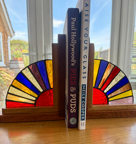 Two wooden bookends with stained-glass fan designs in red, yellow, blue, and green, holding up two books on a wooden table in front of a window.