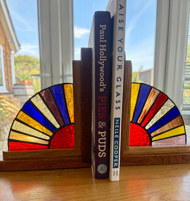 Two wooden bookends with stained-glass fan designs in red, yellow, blue, and green, holding up two books on a wooden table in front of a window.