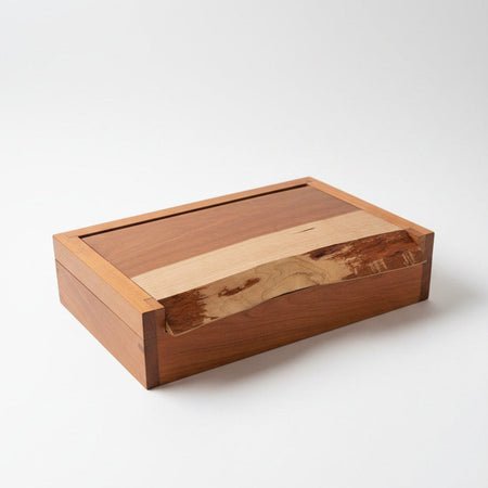 A rectangular wooden box with a hinged lid, featuring a dark reddish-brown cherry wood body and a lighter natural wood grain accent on the front, set against a plain light gray background.