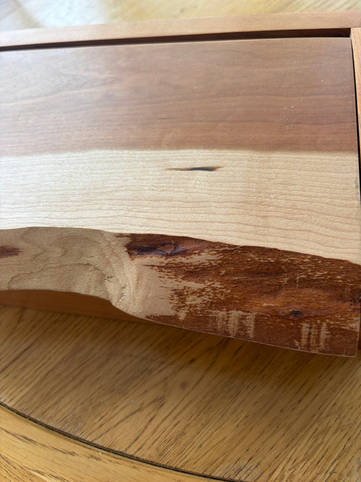 A close-up of a live-edge wood slab showing a natural transition from pale beige wood on the left to medium brown and dark reddish-brown on the right, set against a background of a similar wood grain surface.
