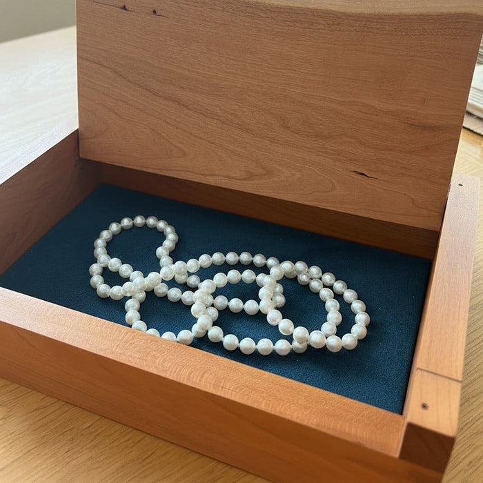 A wooden jewelry box with its lid propped open, featuring a dark blue velvet interior lining and a white beaded necklace resting inside.