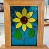 A square stained glass window panel featuring a yellow flower with a brown center and green leaves on a blue background, set in a light oak wood frame.