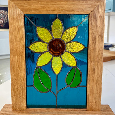 A square stained glass window panel featuring a yellow flower with a brown center and green leaves on a blue background, set in a light oak wood frame.