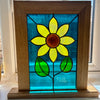 A stained glass sunflower wall art piece in a square wooden frame, displayed on a windowsill. The design features a yellow sunflower with a brown center and green leaves on a textured blue glass background.