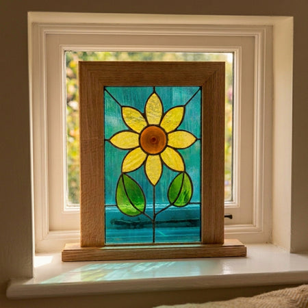 A square stained glass window panel featuring a bright yellow-green sunflower with a brown center, set in a natural oak wood frame. The panel is displayed outdoors on a wooden surface with a blurred garden background.