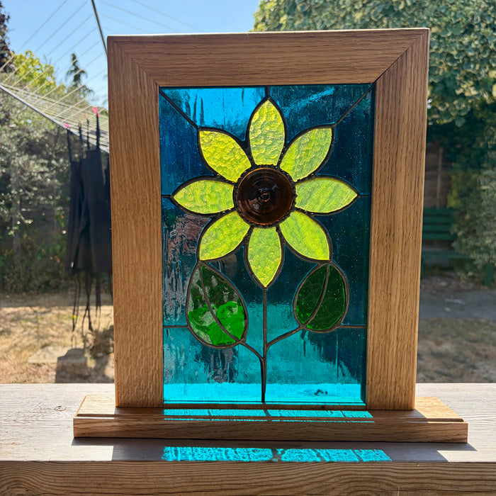 A square stained glass window panel featuring a bright yellow-green sunflower with a brown center, set in a natural oak wood frame. The panel is displayed outdoors on a wooden surface with a blurred garden background.