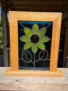 A framed stained glass panel featuring a flower design with green petals, a dark center, and a light wood frame, displayed on a wooden surface.