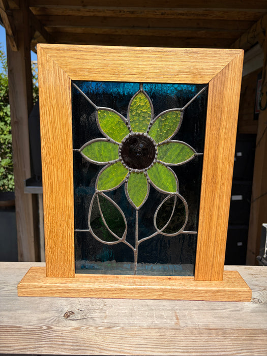 A framed stained glass panel featuring a flower design with green petals, a dark center, and a light wood frame, displayed on a wooden surface.
