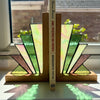 A pair of stained-glass bookends made from fan-shaped pieces in green, cream, pink, and brown with black outlines, resting on a windowsill with a book standing between them.