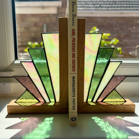 A pair of stained-glass bookends made from fan-shaped pieces in green, cream, pink, and brown with black outlines, resting on a windowsill with a book standing between them.