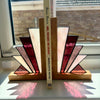 A pair of stained glass bookends shaped like open books, standing on a windowsill. The bookends feature geometric stained glass panels in burgundy, cream, and textured pink, set in a natural oak base.