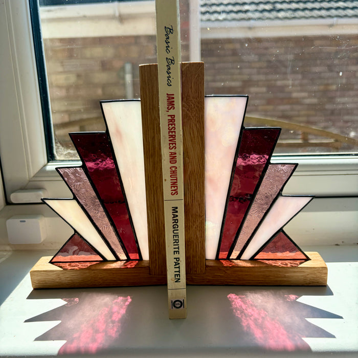 A pair of stained glass bookends shaped like open books, standing on a windowsill. The bookends feature geometric stained glass panels in burgundy, cream, and textured pink, set in a natural oak base.