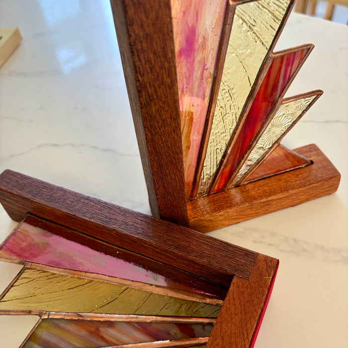 A close-up of a stained glass art piece featuring geometric triangular panels in gold, pink, red, and brown tones, set in a wooden frame on a light marble surface.